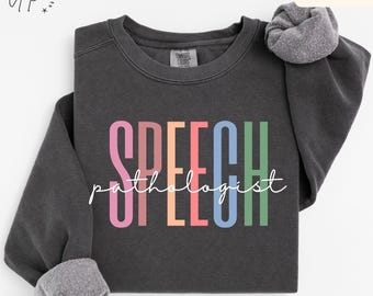 May include: A charcoal gray crewneck sweatshirt featuring the word SPEECH in large, multicolored block letters and the word pathologist written in white cursive script across the center. The garment has long sleeves and a ribbed collar.
