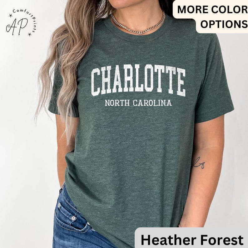 Op de afbeelding: Een donkergroen t-shirt met het woord "CHARLOTTE" in witte blokletters boven "NORTH CAROLINA". Het shirt heeft een ronde hals en korte mouwen.
