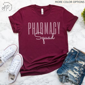 May include: A maroon t-shirt with the text "Pharmacy Squad" in white lettering. The shirt is laid flat on a white surface with a pair of white sneakers and denim shorts.