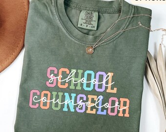 May include: An olive green t-shirt with the words "SCHOOL COUNSELOR" in colourful block letters. The shirt has a crew neck and short sleeves. A silver necklace with a small pendant is draped across the shirt. The shirt is labelled "Comfort Colors".