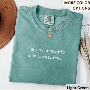May include: A light green t-shirt with the slogan "i'm not dramatic i'm theatrical" in white. The shirt has a crew neck and short sleeves. A gold necklace with a pendant is draped on the shirt.