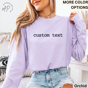 May include: A long-sleeved, light purple shirt with the words "custom text" printed in black across the chest. The shirt has a crew neck and is made of a soft material. The text "MORE COLOR OPTIONS" is visible in the top right corner.