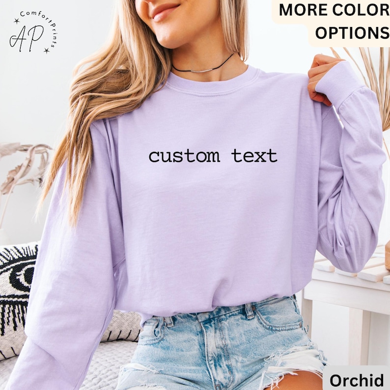 May include: A long-sleeved, light purple shirt with the words "custom text" printed in black across the chest. The shirt has a crew neck and is made of a soft material. The text "MORE COLOR OPTIONS" is visible in the top right corner.