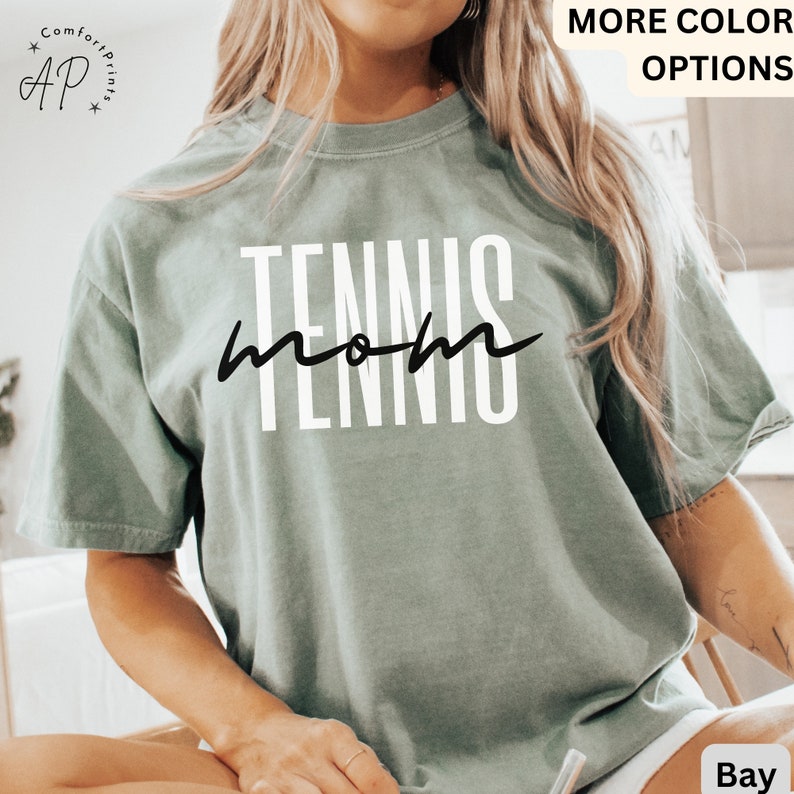 May include: A light green t-shirt with the text "TENNIS mom" in white lettering with black script. The shirt is a relaxed fit and has a crew neck.