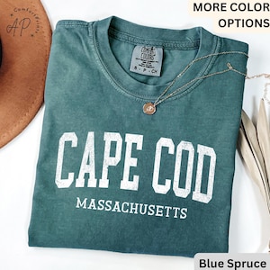 May include: A blue spruce t-shirt with the words "CAPE COD MASSACHUSETTS" printed in white block letters. The shirt has a crew neck and short sleeves. A gold necklace with a pendant is draped across the shirt.