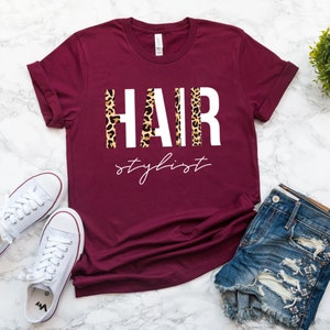 May include: Burgundy t-shirt with the words "HAIR stylist" in white letters with a leopard print pattern behind the letters.