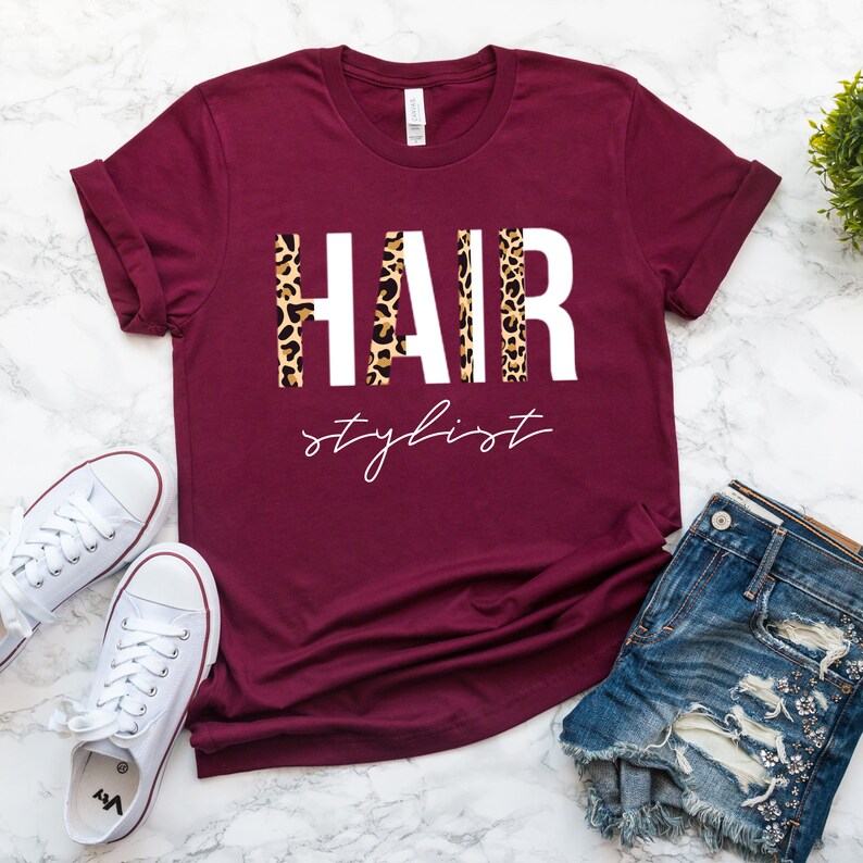 May include: Burgundy t-shirt with the words "HAIR stylist" in white letters with a leopard print pattern behind the letters.