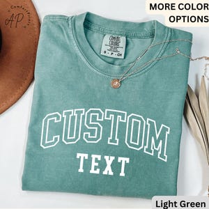 Puede incluir: Camiseta verde claro con texto blanco que dice "CUSTOM TEXT" en una fuente universitaria.