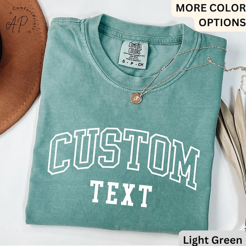 Puede incluir: Camiseta verde claro con texto blanco que dice "CUSTOM TEXT" en una fuente universitaria.