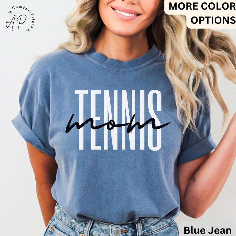 May include: A blue jean t-shirt with white text that reads "Tennis Mom" in a modern font. The shirt is being worn by a person with long blonde hair.