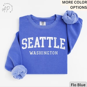 May include: Flo Blue sweatshirt with the word "SEATTLE" and "WASHINGTON" printed in white block letters. The sweatshirt has a crew neck and long sleeves. The sleeves are cuffed. The sweatshirt is made of a soft material.