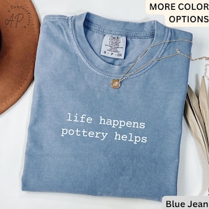 May include: A blue jean coloured t-shirt with the text "life happens pottery helps" in white. The shirt has a crew neck and short sleeves. A gold necklace with a pendant is draped on the shirt.