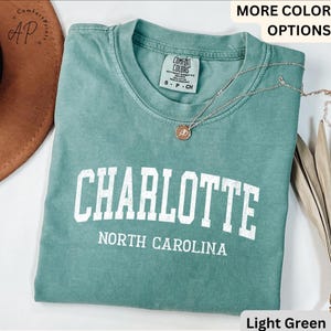 Op de afbeelding: Lichtgroen t-shirt met het woord "CHARLOTTE" in witte blokletters boven "NORTH CAROLINA". Het shirt heeft een ronde hals en korte mouwen. Een zilveren ketting met een hanger is over het shirt gedrapeerd.