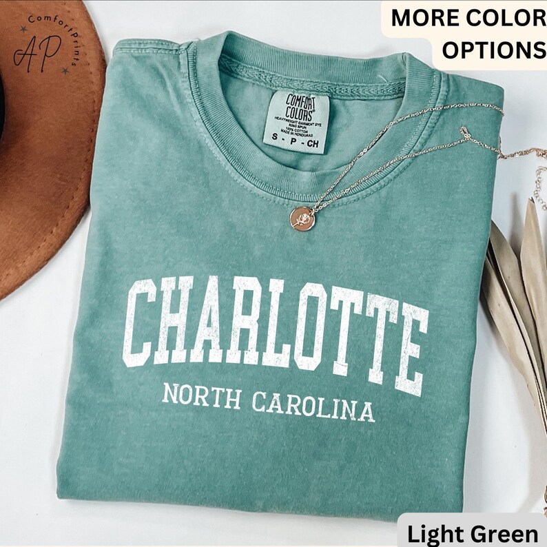 Op de afbeelding: Lichtgroen t-shirt met het woord "CHARLOTTE" in witte blokletters boven "NORTH CAROLINA". Het shirt heeft een ronde hals en korte mouwen. Een zilveren ketting met een hanger is over het shirt gedrapeerd.