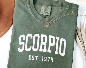 Pode incluir: Uma camiseta verde musgo com a palavra "SCORPIO" em letras brancas e "EST. 1974" abaixo. Um colar de prata com um pequeno pingente repousa sobre a camisa. A camisa está dobrada e um chapéu marrom é parcialmente visível.