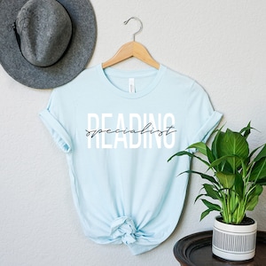 Puede incluir: Una camiseta azul claro con el texto "READING specialist" en letras blancas con un "specialist" en cursiva negra en una percha.