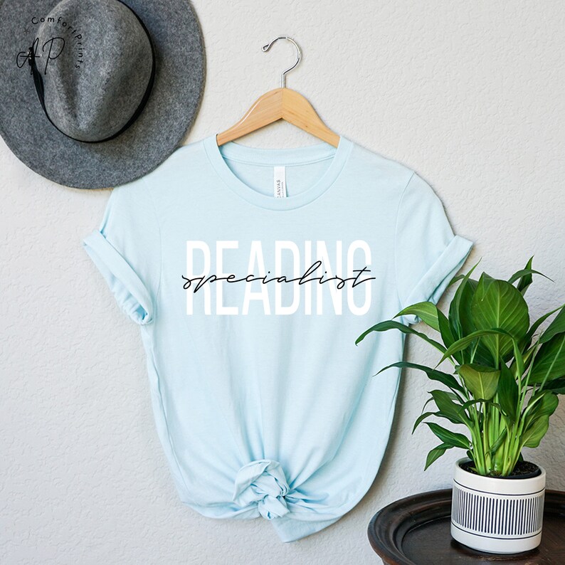 Puede incluir: Una camiseta azul claro con el texto "READING specialist" en letras blancas con un "specialist" en cursiva negra en una percha.