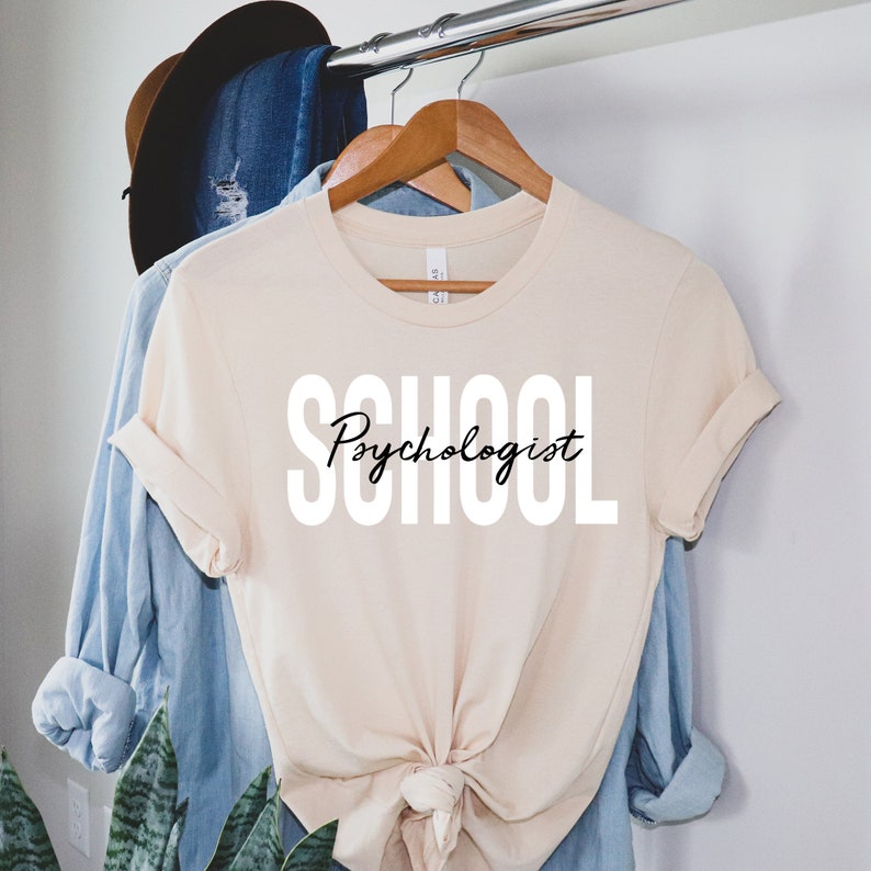 Op de afbeelding: Een beige T-shirt met de tekst "School Psychologist" in witte letters met zwarte letters voor het woord "Psychologist".