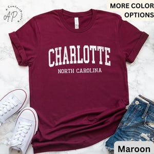 Op de afbeelding: Bordeauxrood t-shirt met het woord "CHARLOTTE" in witte blokletters boven "NORTH CAROLINA". Het shirt wordt getoond met witte sneakers en een spijkerbroekshort. De woorden "MORE COLOR OPTIONS" staan bovenaan.