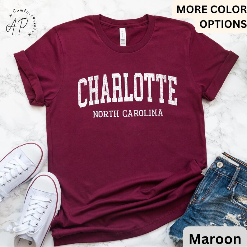 Op de afbeelding: Bordeauxrood t-shirt met het woord "CHARLOTTE" in witte blokletters boven "NORTH CAROLINA". Het shirt wordt getoond met witte sneakers en een spijkerbroekshort. De woorden "MORE COLOR OPTIONS" staan bovenaan.