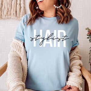 K&ouml;nnte beinhalten: Ein hellblaues T-Shirt mit dem Aufdruck "Hair stylist" in Wei&szlig; auf der Vorderseite.