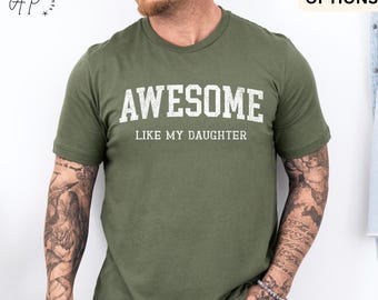 May include: Military green t-shirt with the word "AWESOME" in white block letters above the phrase "LIKE MY DAUGHTER". The shirt has a crew neck and short sleeves. The text is slightly distressed.
