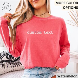 May include: A watermelon-colored long-sleeve sweatshirt with the words "custom text" in white. The sweatshirt has a crew neck and ribbed cuffs. The model is wearing denim shorts. The brand logo "AP" is visible in the upper left corner.