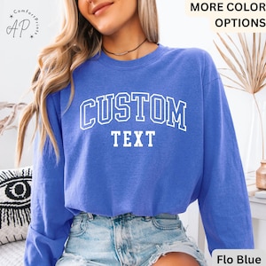 May include: A blue sweatshirt with the words "CUSTOM TEXT" in white block letters. The sweatshirt has a crew neck and long sleeves. The text is in a collegiate style font. The words "MORE COLOR OPTIONS" are in the top right corner.