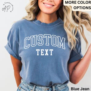 Puede incluir: Una camiseta azul jean con texto blanco que dice "CUSTOM TEXT" en una fuente universitaria.