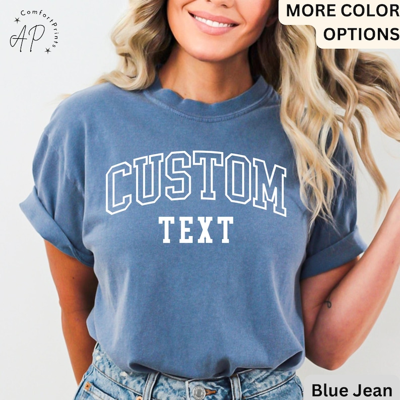 Puede incluir: Una camiseta azul jean con texto blanco que dice "CUSTOM TEXT" en una fuente universitaria.