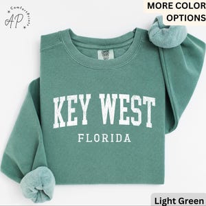 May include: Light green sweatshirt with the text "KEY WEST FLORIDA" in white block letters. The sweatshirt has a crew neck and long sleeves with a light green, soft, fuzzy lining. The words "MORE COLOR OPTIONS" are at the top.