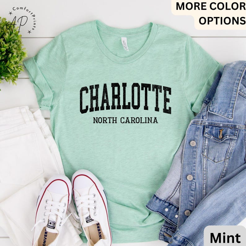 Op de afbeelding: Een mintgroen t-shirt met het woord "CHARLOTTE" in vette zwarte letters boven "NORTH CAROLINA". Het shirt wordt gecombineerd met witte sneakers, een witte jeans en een spijkerjasje. Het shirt heeft een casual stijl.