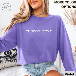May include: A violet long-sleeve sweatshirt with the text "custom text" in white. The sweatshirt is a solid color and has a relaxed fit. The image also includes the words "MORE COLOR OPTIONS" and "Violet".