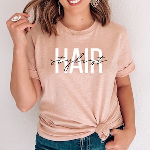 K&ouml;nnte beinhalten: Ein hellrosa T-Shirt mit dem Aufdruck "HAIR stylist" in Wei&szlig; mit schwarzer Schrift.
