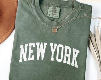 May include: A moss green t-shirt with the text "NEW YORK" in white block letters. The shirt has a crew neck and short sleeves. A silver necklace with a small pendant is draped across the shirt. The shirt is folded and appears to be made of a soft material.