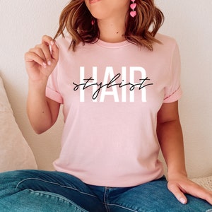 K&ouml;nnte beinhalten: Ein pinkfarbenes T-Shirt mit dem Text "HAIR stylist" in wei&szlig;en Buchstaben mit schwarzem Umriss.