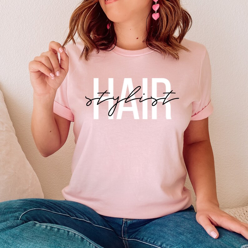 K&ouml;nnte beinhalten: Ein pinkfarbenes T-Shirt mit dem Text "HAIR stylist" in wei&szlig;en Buchstaben mit schwarzem Umriss.