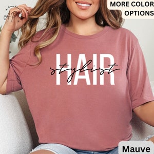 K&ouml;nnte beinhalten: Mauvefarbenes T-Shirt mit dem Text "Hair Stylist" in wei&szlig;en Buchstaben.