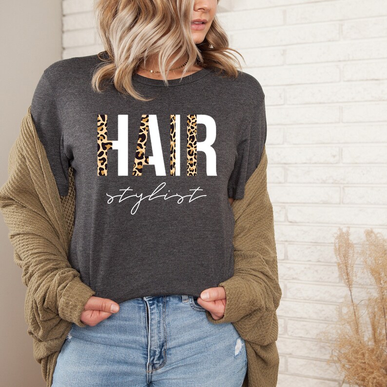 May include: A dark gray t-shirt with the text "HAIR stylist" in white letters. The word "HAIR" is made up of leopard print.