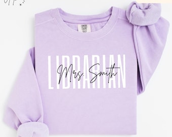 May include: A light purple sweatshirt with the word "LIBRARIAN" in white block letters and "Mrs. Smith" in cursive. The sweatshirt has long sleeves and a crew neck. The color is described as "Orchid".