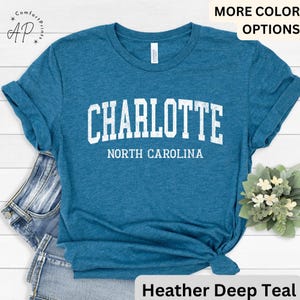 Op de afbeelding: Tealkleurig T-shirt met het woord "CHARLOTTE" in witte blokletters boven "NORTH CAROLINA". Het shirt heeft een heather deep teal kleur en heeft opgerolde mouwen. Het shirt is aan de onderkant geknoopt.