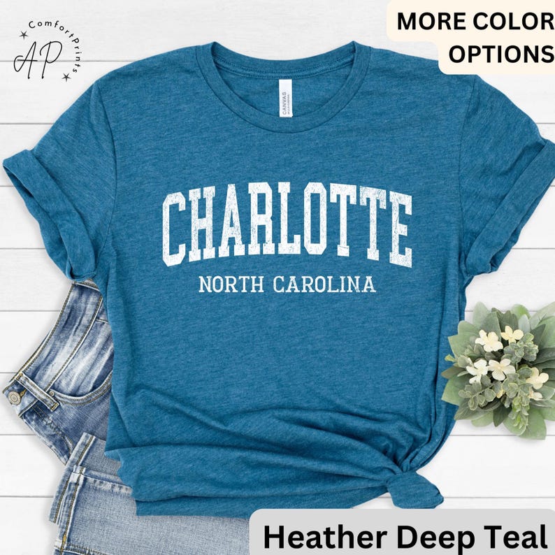 Op de afbeelding: Tealkleurig T-shirt met het woord "CHARLOTTE" in witte blokletters boven "NORTH CAROLINA". Het shirt heeft een heather deep teal kleur en heeft opgerolde mouwen. Het shirt is aan de onderkant geknoopt.