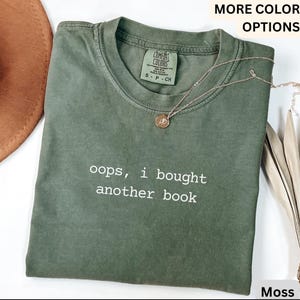 May include: A moss green t-shirt with the text "oops, i bought another book" in white. The shirt has a crew neck and short sleeves. A gold necklace with a small pendant rests on the shirt. The shirt is folded.
