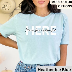 Puede incluir: Camiseta azul hielo con el texto "HERE Place Your Custom Text". La camiseta tiene mangas cortas y cuello redondo. Las palabras "MORE COLOR OPTIONS" est&aacute;n en la esquina superior derecha. El logotipo de la marca "AP" est&aacute; en la esquina superior izquierda.