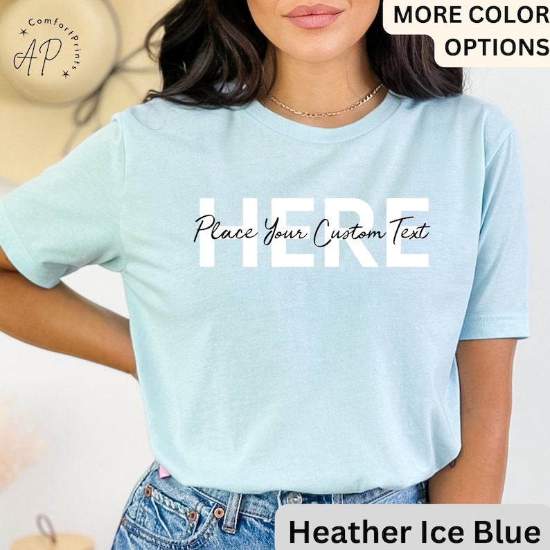 Puede incluir: Camiseta azul hielo con el texto "HERE Place Your Custom Text". La camiseta tiene mangas cortas y cuello redondo. Las palabras "MORE COLOR OPTIONS" est&aacute;n en la esquina superior derecha. El logotipo de la marca "AP" est&aacute; en la esquina superior izquierda.