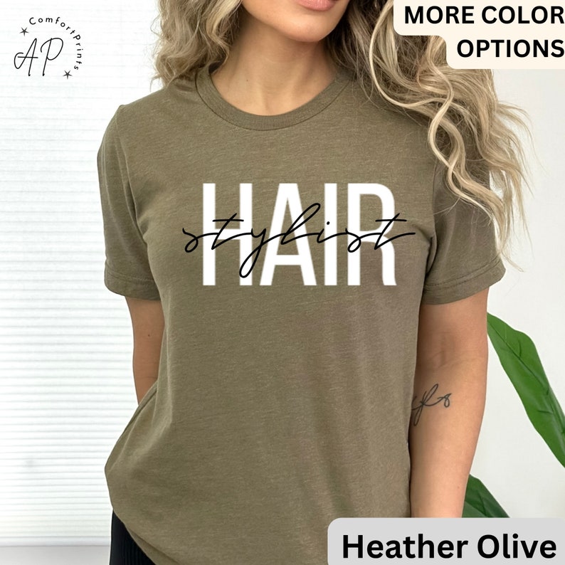 K&ouml;nnte beinhalten: Ein gr&uuml;nes T-Shirt mit dem Text "Hair Stylist" in wei&szlig;en Buchstaben mit schwarzer kursivschrift.