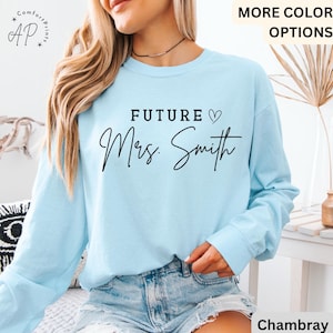 May include: A light blue long-sleeve shirt with the text "FUTURE Mrs. Smith" in black script. The shirt has a crew neck and is made of a soft material. The shirt is a bridal shower or wedding gift.