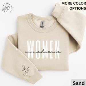 May include: A sand-coloured sweatshirt with the text "WOMEN in medicine" printed in white on the front. The left sleeve has a small black stethoscope graphic with the letters "AP" underneath.