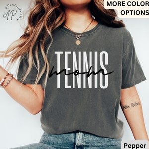 May include: A gray t-shirt with the text "Tennis Mom" in white and black lettering. The shirt is worn by a person with long brown hair.