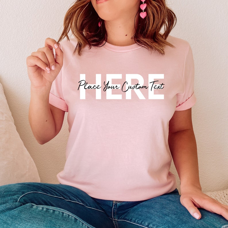 Puede incluir: Una camiseta rosa claro con mangas enrolladas. La camiseta presenta la palabra "HERE" en letras may&uacute;sculas blancas grandes, con la frase "Place Your Custom Text" debajo en una fuente cursiva m&aacute;s peque&ntilde;a. El modelo lleva vaqueros azules.